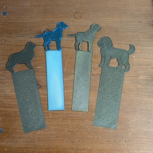 Pode incluir: Quatro marcadores de página com silhuetas de cães no topo. O primeiro é verde-escuro, o segundo tem um cão azul sobre uma base retangular azul-clara, o terceiro é verde metálico e o quarto é um cão verde metálico com acabamento texturizado.