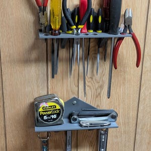 Puede incluir: Un organizador de herramientas gris de pared con una variedad de herramientas, incluyendo alicates, destornilladores, una cinta métrica, una llave y un calibre. El organizador tiene un estante de metal gris y una base de plástico gris.