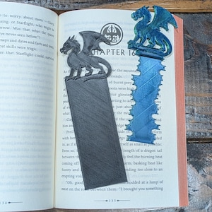Pode incluir: Dois marcadores em forma de dragão estão nas páginas de um livro aberto. Um é cinzento, o outro azul e verde. O livro está aberto no Capítulo 16. Os marcadores são feitos de um material texturizado.