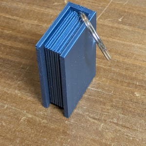 Pode incluir: Um pequeno livro em miniatura azul impresso em 3D com um porta-chaves prateado. O livro tem bordas de página detalhadas e uma forma retangular. O porta-chaves está preso ao topo do livro. O item está em uma superfície de madeira.