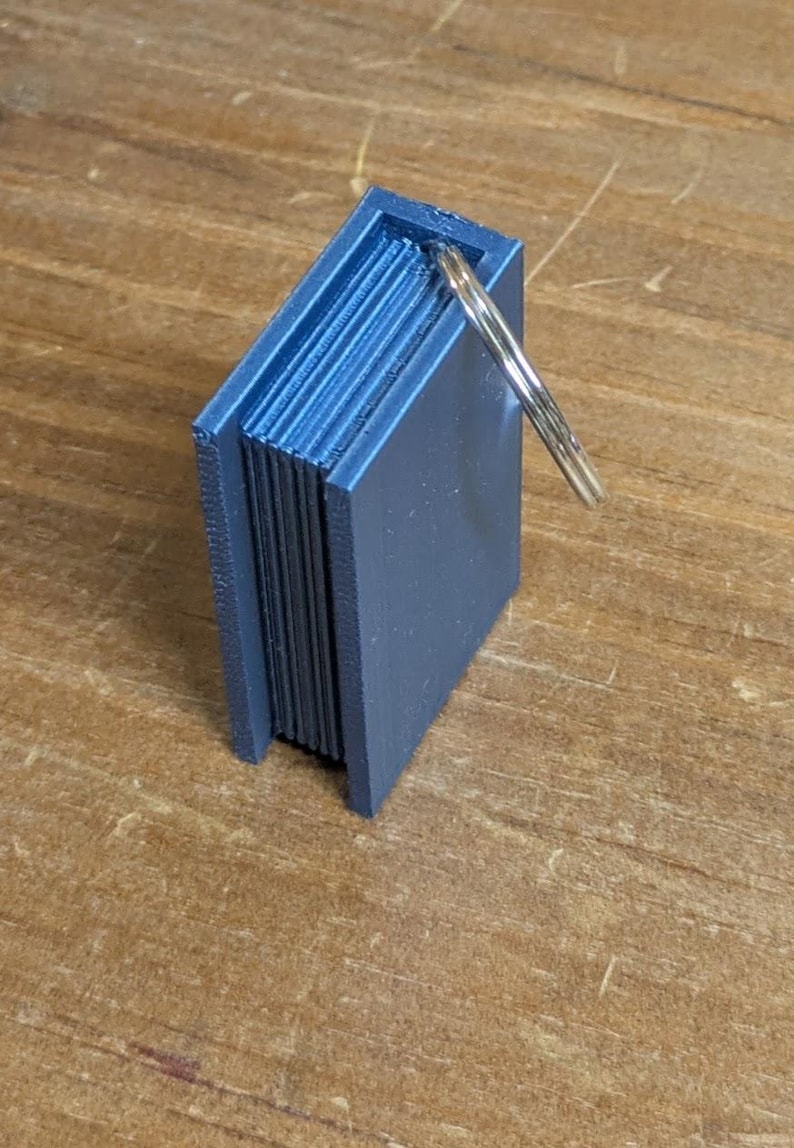May include: A small, blue, 3D-printed miniature book with a silver keyring attached. The book has detailed page edges and a rectangular shape. The keyring is attached to the top of the book. The item is sitting on a wooden surface.