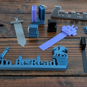 Könnte beinhalten: Eine Sammlung von 3D-gedruckten Lesezeichen und Buchzubehör in verschiedenen Formen und Farben. Enthält Schwerter, Gitarren und Buchdesigns sowie ein graues Lesezeichen mit der Aufschrift "To Be Read".