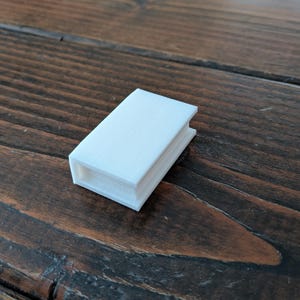 May include: A small, white, rectangular object with a book-like appearance. The object has a textured surface and appears to be 3D-printed. It is resting on a dark brown wooden surface.