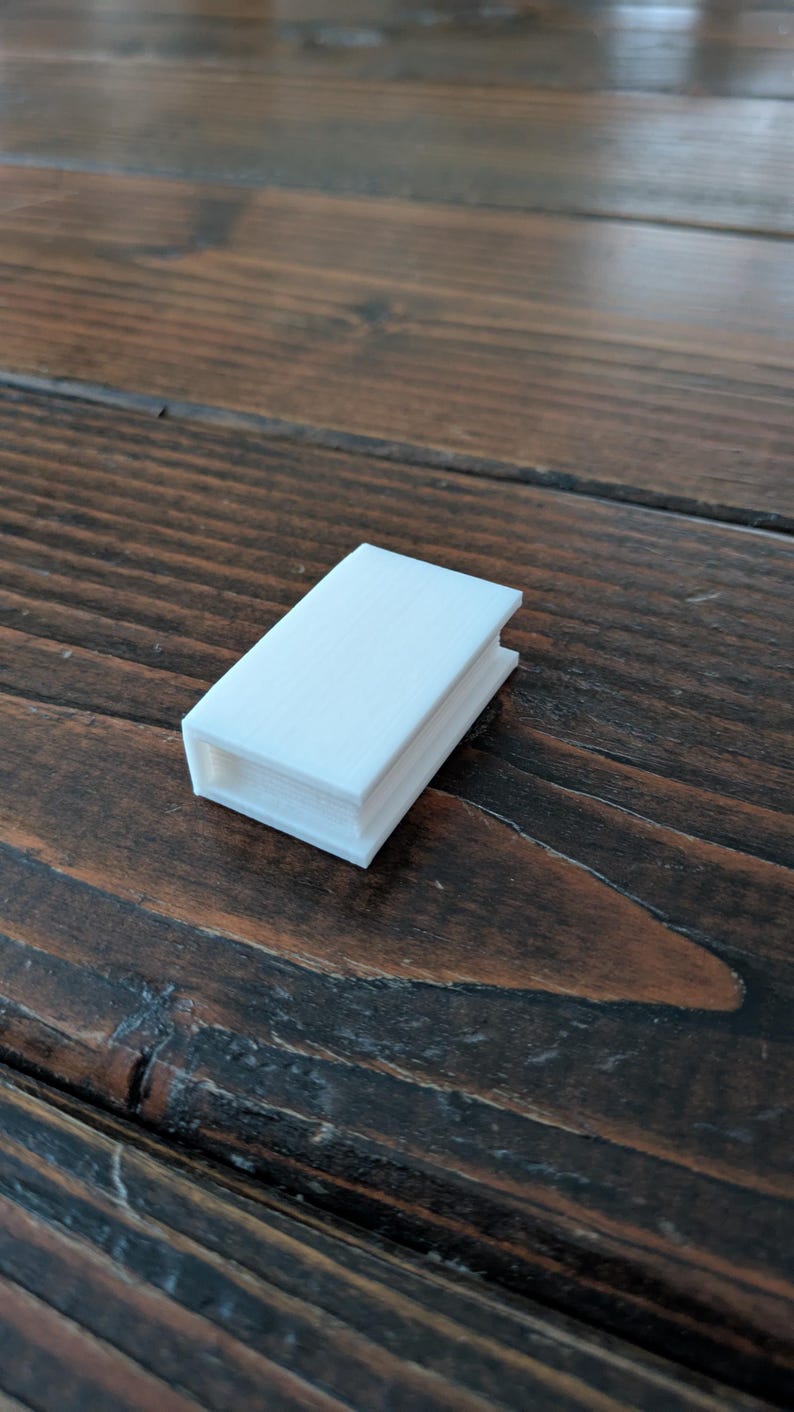 May include: A small, white, rectangular object with a book-like appearance. The object has a textured surface and appears to be 3D-printed. It is resting on a dark brown wooden surface.