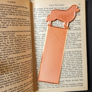 May include: A close-up of an open book with a brown bookmark shaped like a dog. The book is titled "I, Robot" and the bookmark is titled "Catch That Rabbit". The text on the page discusses robots and their potential for danger.