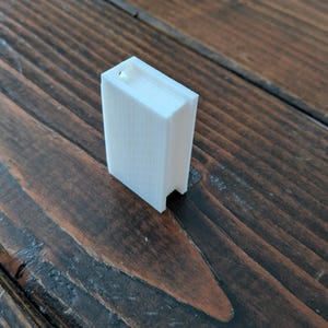 May include: A small, white, rectangular 3D-printed object resembling a book. The object has a slot at the top and is positioned on a dark brown wooden surface. The object's edges are clearly defined, and the overall design is minimalist.