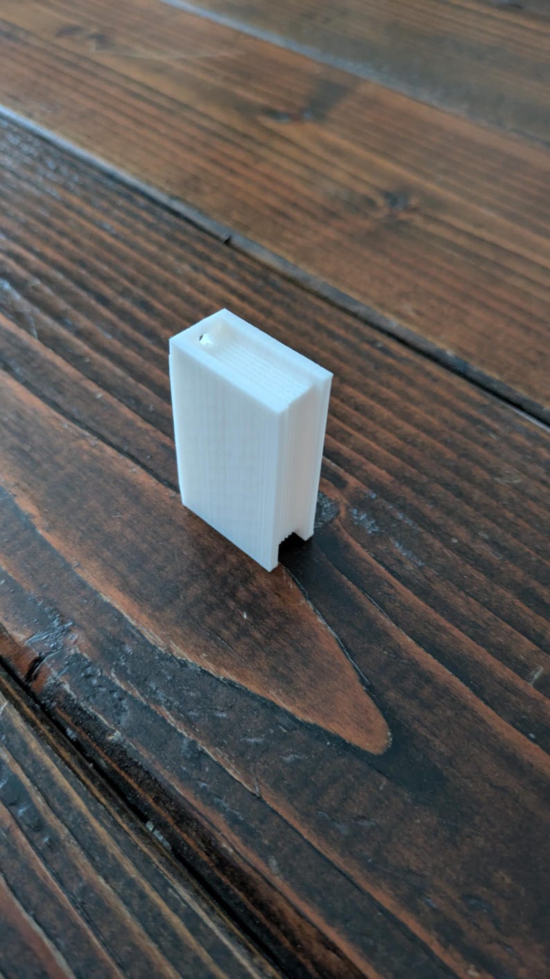 May include: A small, white, rectangular 3D-printed object resembling a book. The object has a slot at the top and is positioned on a dark brown wooden surface. The object's edges are clearly defined, and the overall design is minimalist.
