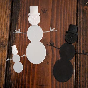 Pode incluir: Três decorações de boneco de neve em branco e preto, cada uma com um chapéu alto e braços de pau. Os bonecos de neve estão dispostos sobre uma superfície de madeira escura. O boneco de neve maior é branco, o menor é branco e o terceiro é preto.