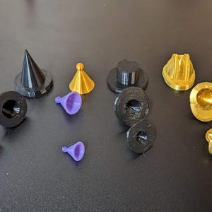 Peut inclure: Assortiment de petits objets imprimés en 3D de différentes couleurs, notamment noir, or et violet. Les objets sont en forme de cône, cylindriques et présentent une variété de motifs.