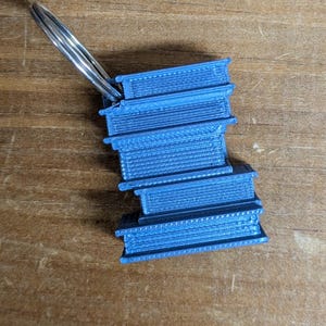 May include: A blue 3D-printed keychain shaped like a stack of books. The keychain is attached to a silver metal ring. The books have detailed textures and are stacked unevenly. The background is a wooden surface.