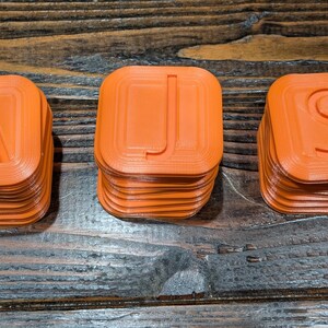 May include: Three orange plastic blocks stacked on top of each other, spelling out the word "AJS".