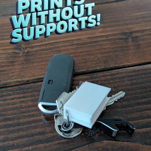 以下が含まれることがあります： 木製の表面に置かれた、白い3Dプリントされた本のキーホルダー。キーホルダーは、鍵、黒い車のキーホルダー、黒い栓抜きに繋がっています。画像には「PRINTS WITHOUT SUPPORTS!」というテキストが表示されています。