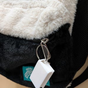 May include: A plush backpack with a cat face design, featuring white and black fur. The backpack has a zipper and a small white tag attached. The tag is rectangular and appears to be a keychain.