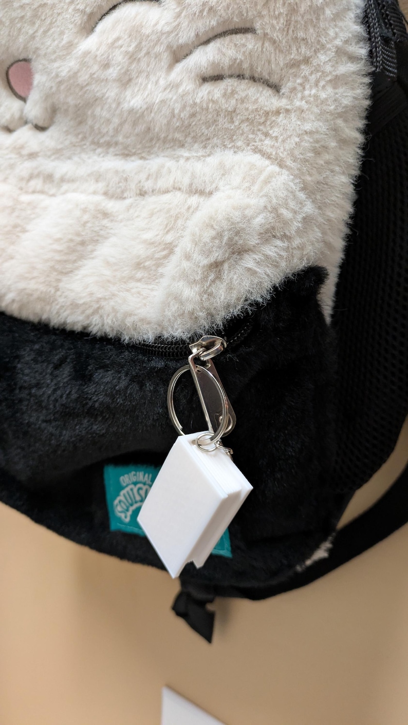May include: A plush backpack with a cat face design, featuring white and black fur. The backpack has a zipper and a small white tag attached. The tag is rectangular and appears to be a keychain.