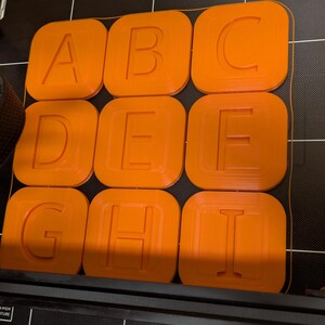 May include: Nine orange 3D printed squares with the letters A, B, C, D, E, F, G, H, and I printed in white on each square.