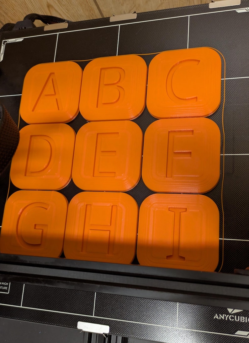 May include: Nine orange 3D printed squares with the letters A, B, C, D, E, F, G, H, and I printed in white on each square.