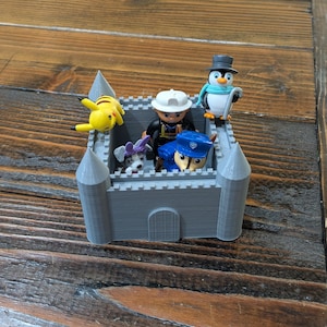 Op de afbeelding: Een grijs plastic kasteel met een gele Pikachu, een pinguïn met een blauwe sjaal, een hond met paarse oren en een persoon met een zwarte hoed en een blauw shirt erin.