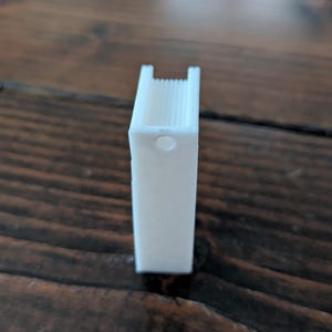 May include: A white, rectangular 3D-printed object with a hole and a ridged top. The object is standing upright on a dark brown wooden surface. The object appears to be a small binder clip.