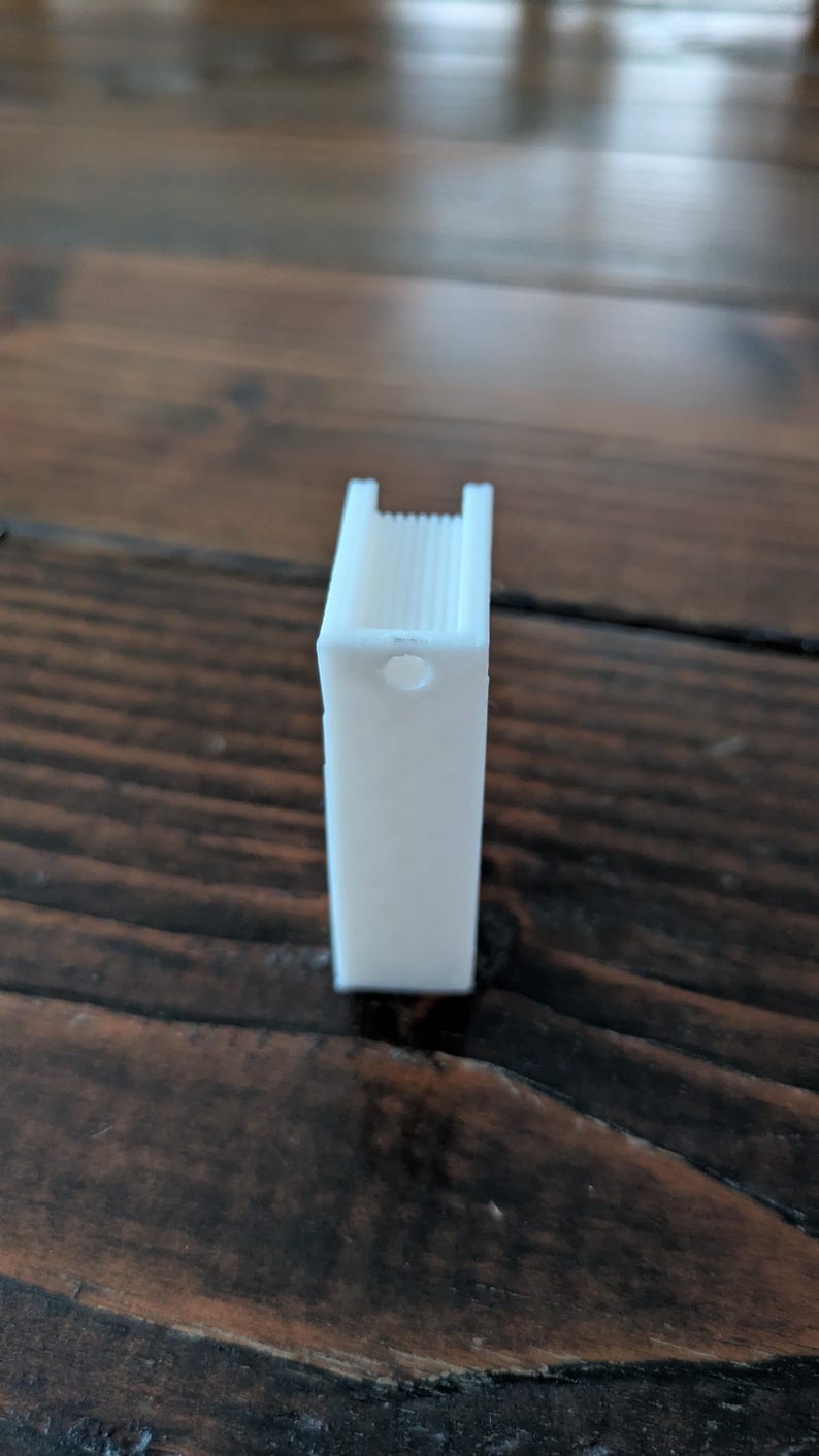 May include: A white, rectangular 3D-printed object with a hole and a ridged top. The object is standing upright on a dark brown wooden surface. The object appears to be a small binder clip.