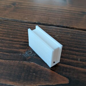 May include: A small, white, rectangular object with a ridged top and a hollowed-out interior. The object is made of a solid material and sits on a dark wooden surface. The object appears to be a small container or holder.
