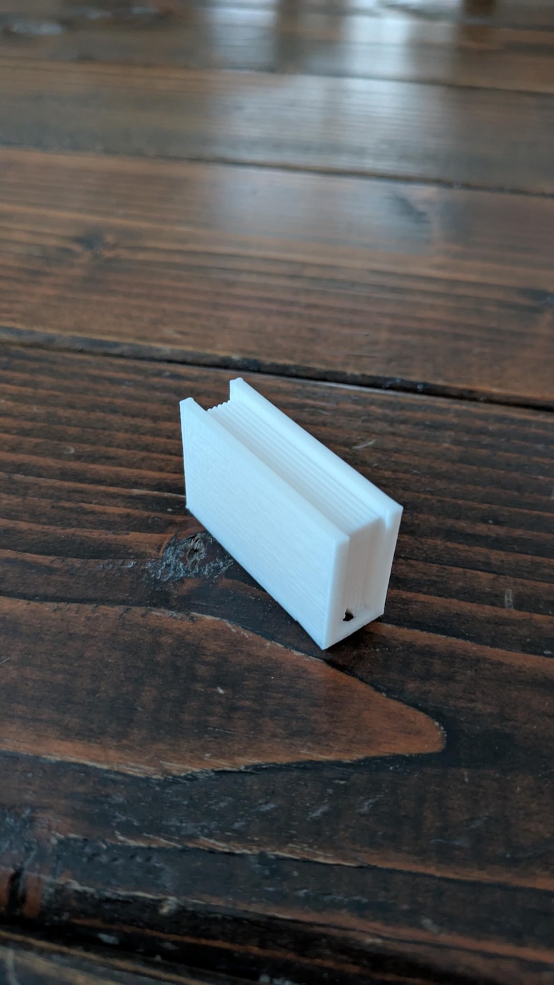 May include: A small, white, rectangular object with a ridged top and a hollowed-out interior. The object is made of a solid material and sits on a dark wooden surface. The object appears to be a small container or holder.