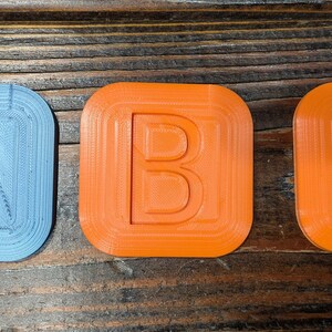 May include: Three 3D printed plastic squares, each with a different letter of the alphabet. The letter A is blue, the letter B is orange, and the letter C is orange.
