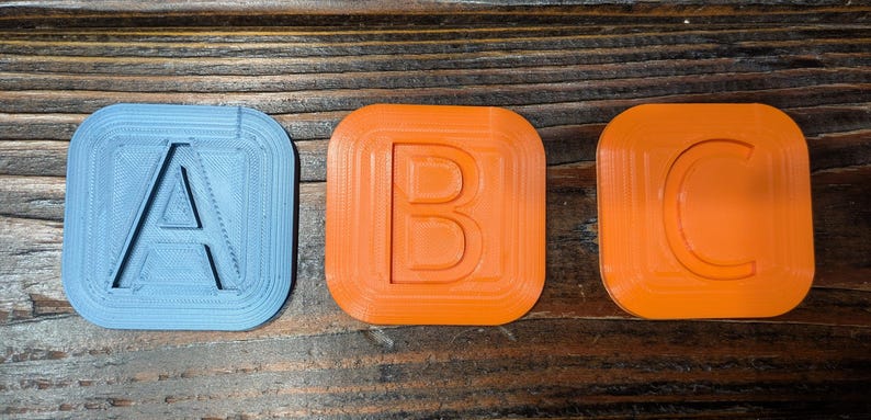 May include: Three 3D printed plastic squares, each with a different letter of the alphabet. The letter A is blue, the letter B is orange, and the letter C is orange.
