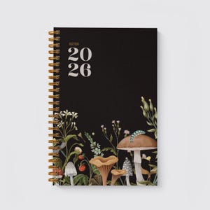 May include: Black spiral-bound agenda for 2026 with gold-coloured binding. The cover features the word "AGENDA" above the numbers "20 26" in white. A botanical illustration of mushrooms, flowers, and foliage decorates the bottom edge.
