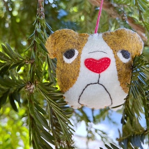 May include: A felt ornament shaped like a bulldog with brown ears and a white face. The bulldog has a pink heart nose and black eyes.