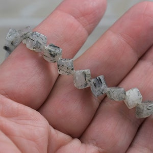Può includere: Una collana di perle di quarzo trasparente sfaccettate con inclusioni nere. Le perle sono infilate su un filo elastico trasparente e hanno un diametro di circa 8 mm.