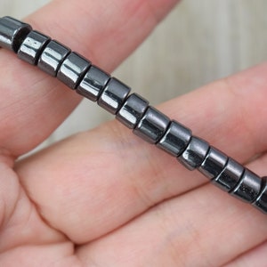 May include: A strand of dark grey, rectangular hematite beads. The beads are strung together, forming a flexible chain. The beads have a metallic sheen and are held in a hand.