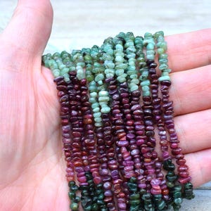 May include: A strand of green and red gemstone beads. The beads are small and irregular in shape, and they are strung on a clear thread. The beads are arranged in a gradient pattern, with the green beads at the top and the red beads at the bottom.