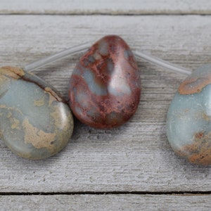 May include: Three teardrop-shaped gemstone beads on a clear string. The beads display a mix of colours, including blue-grey, brown, and reddish-brown. The beads are smooth and polished, with natural patterns. The background is a light-coloured wooden surface.