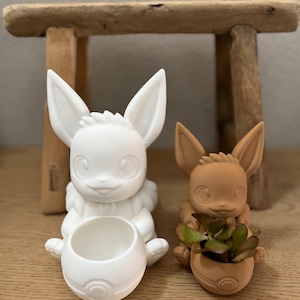 Puede incluir: Dos macetas de cerámica con forma de personaje Pokémon, Eevee. La maceta de la izquierda es blanca y la de la derecha es marrón. La maceta marrón tiene una pequeña planta suculenta en su interior.