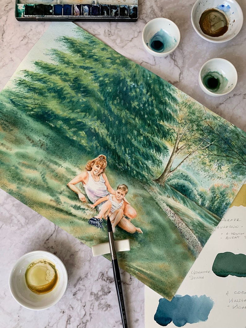May include: A watercolor painting of a mother and child sitting in a grassy field. The woman is wearing a white tank top and the child is wearing a blue shirt. There are trees in the background and a small stream running through the field. The painting is on a white background and there are paintbrushes and paint palettes visible in the foreground.