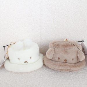 May include: Two plush hats, one white and one tan, each with a cartoon face and a small brim. The white hat has bunny ears, while the tan hat has bear ears. Both have a gold chain and a tag.