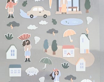May include: Sheet of Suatelier stickers featuring rainy day illustrations. Includes images of people with umbrellas, houses, cars, clouds, and a bus stop. The stickers are in various colors, including blue, green, and orange.
