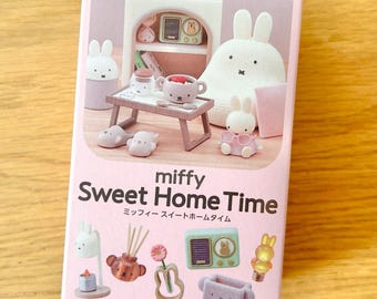 May include: A rectangular pink product box for a Miffy Sweet Home Time miniature set. The packaging features illustrations of various Miffy-themed furniture and accessories, including a small table, a radio, and several Miffy character figures.