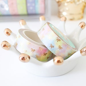 May include: Pastel washi tape rolls with gold bow accents rest in a white crown-shaped dish with gold tips. Additional rolls of colorful washi tape are visible in a clear container in the background. The tape has a watercolor effect.