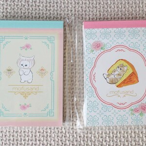 May include: Two small notebooks in plastic packaging. One has a light blue and pink border with a cartoon character. The other has a pink border with a cat and cake illustration. Both notebooks have the text "mofusand".