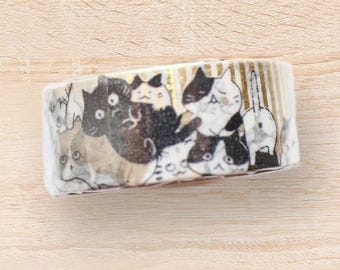 May include: A roll of decorative washi tape featuring a repeating pattern of cartoon cats in various poses and colors, including black, white, and gray. The tape is on a light wood surface.