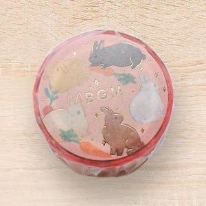 Puede incluir: Rollo de cinta adhesiva decorativa con un diseño rosa que presenta conejos, zanahorias y follaje. La cinta tiene un borde rojo y las letras "MBGM" son visibles.
