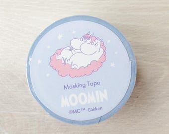 May include: A roll of masking tape featuring a light blue design with a cartoon illustration of Moomin characters. The tape has the text "Masking Tape" and "MOOMIN" printed on it. The tape is on a light wood surface.