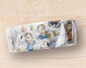 May include: A roll of decorative tape featuring cartoon characters, including Snoopy, Charlie Brown, and Woodstock. The tape has a white background with colorful illustrations of the characters. The tape is packaged in clear plastic and rests on a light wood surface.