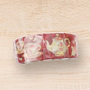 Könnte beinhalten: Rolle dekoratives Washi-Tape mit einem sich wiederholenden Muster aus Teetassen, Teekannen und floralen Designs auf burgunderrotem Hintergrund. Das Klebeband ist in klarem Kunststoff verpackt und liegt auf einer hellen Holzoberfläche.