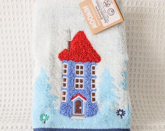 May include: A light blue hand towel featuring a textured, embroidered Moomin house with a bright red roof, a dark blue bottom border, and small floral accents. A tan cardboard tag with the text MOOMIN is attached to the top corner of the towel.