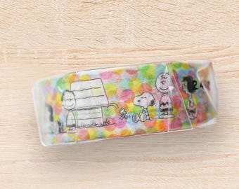May include: A roll of decorative tape featuring Snoopy, Charlie Brown, and other Peanuts characters. The tape has a colorful polka dot pattern and is packaged in clear plastic. The illustrations are in black and white.