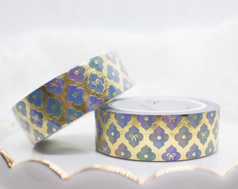 May include: Two rolls of decorative washi tape with a gold, blue, and purple floral pattern. The tape is displayed on a white, scalloped-edge cake stand. The design includes small bow accents.