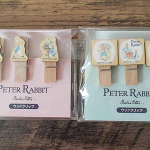 May include: Two sets of Peter Rabbit wooden pegs in clear packaging. One set has three pegs with rabbit illustrations, the other has three square pegs with different Peter Rabbit images. The text "Peter Rabbit" is visible.