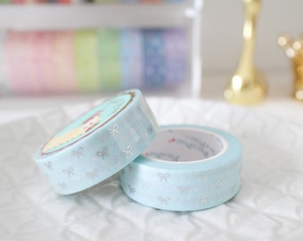May include: Two rolls of decorative washi tape. The tape is light blue with a silver bow pattern. One roll has a circular image of a cartoon character. The tape is on a white, textured surface.